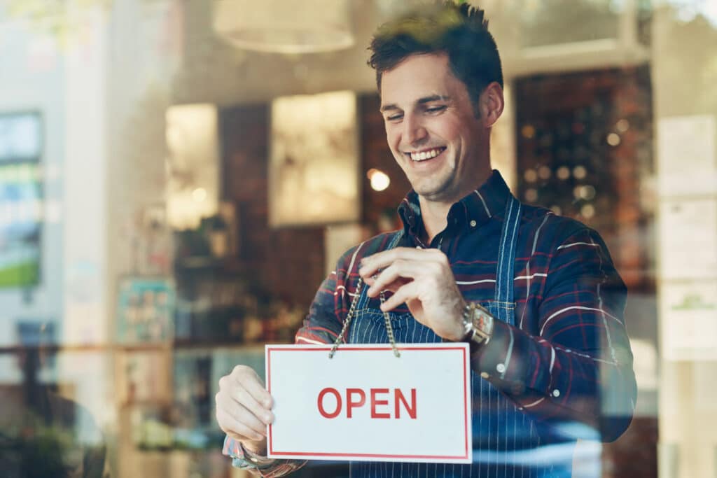 Open sign, cafe and man with business, startup and poster with service, board and franchise window. Male person, happy employee and entrepreneur with a waiter, new and opening store with hospitality.