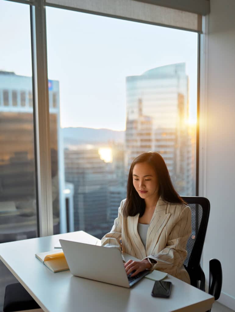 Digital finance professional using her computer at a window overlooking a modern city at sunset